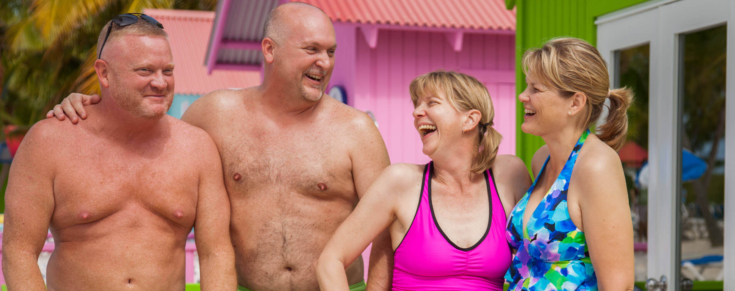 two shirtless men laughing with two women in bathing suits in front of colorful bungalow